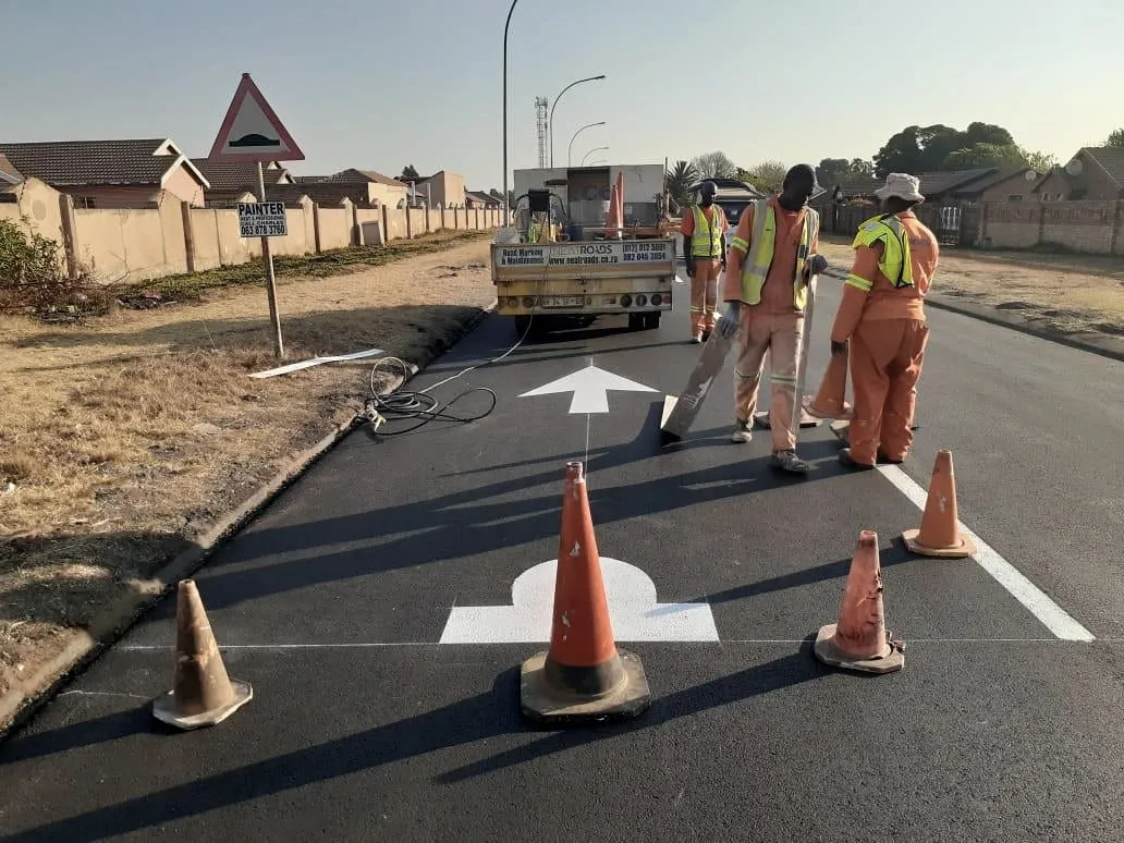 A road-marking crew paints white directional arrows on a freshly resurfaced road.