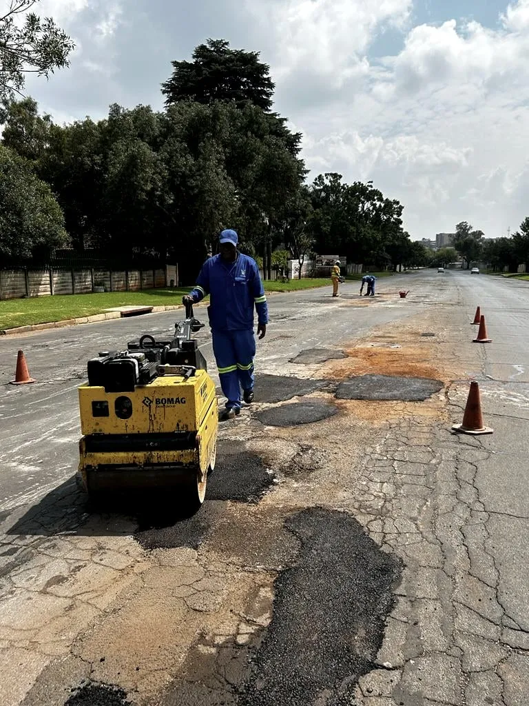 A worker compacts a fresh asphalt patch with a roller on a suburban road.